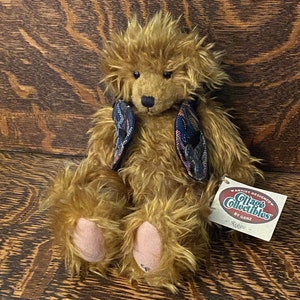 Puede incluir: Un oso de peluche marrón con una textura de pelaje desgreñado, con detalles de tela estampada. El oso tiene almohadillas de patas de color rosa claro y una etiqueta que dice "Cottage Collectibles by Ganz Reggie."