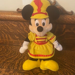 May include: A plush toy of Minnie Mouse dressed as a band member. She is wearing a yellow dress with red trim, a yellow hat with a red feather, and yellow shoes.