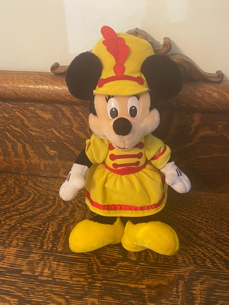May include: A plush toy of Minnie Mouse dressed as a band member. She is wearing a yellow dress with red trim, a yellow hat with a red feather, and yellow shoes.