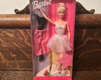 May include: A Barbie doll dressed in a pink and white ballerina costume. The doll is holding a pink rose and is standing on a wooden stage. The box has the text "Barbie Ballerina Ballet Etoile de Ballet" on it.