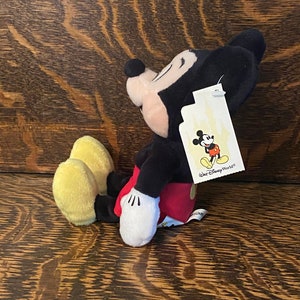 May include: A plush Mickey Mouse toy sitting with its legs crossed. The toy is wearing red shorts and yellow shoes. The toy has a black tag attached to its ear that says "Walt Disney World".