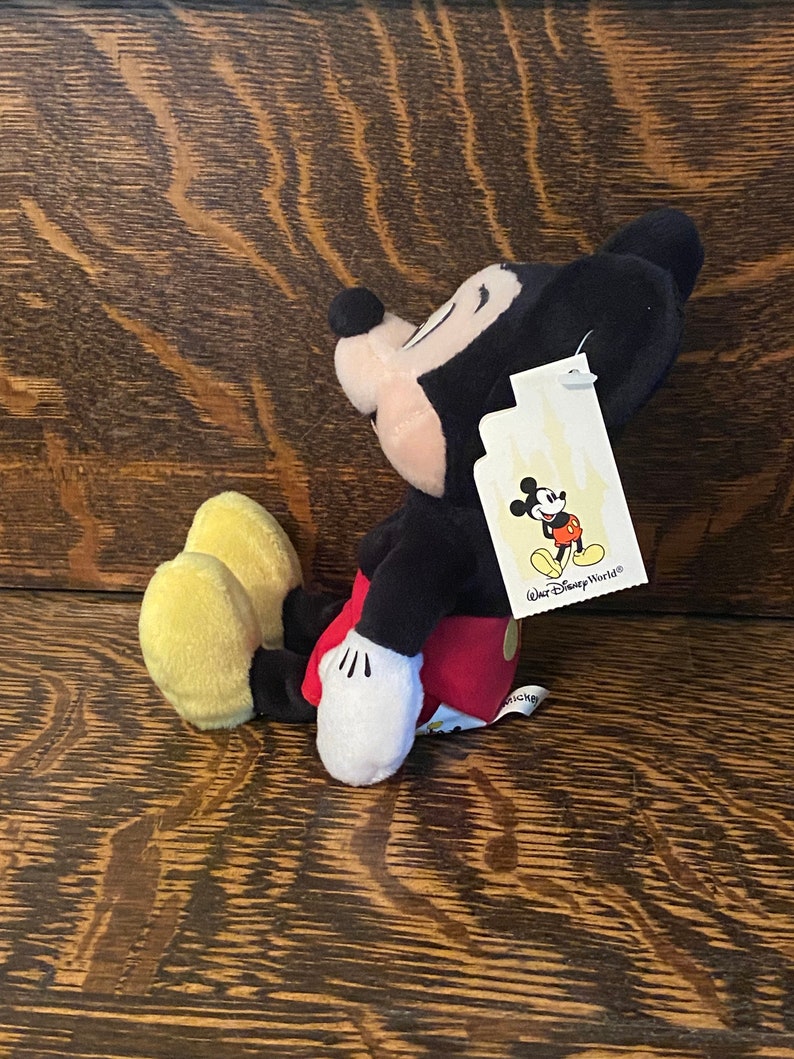 May include: A plush Mickey Mouse toy sitting with its legs crossed. The toy is wearing red shorts and yellow shoes. The toy has a black tag attached to its ear that says "Walt Disney World".