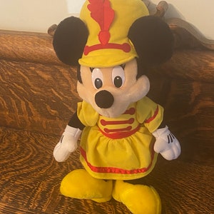 May include: A yellow plush toy of Minnie Mouse dressed as a drum major. She is wearing a yellow dress with red trim and a yellow hat with a red feather.
