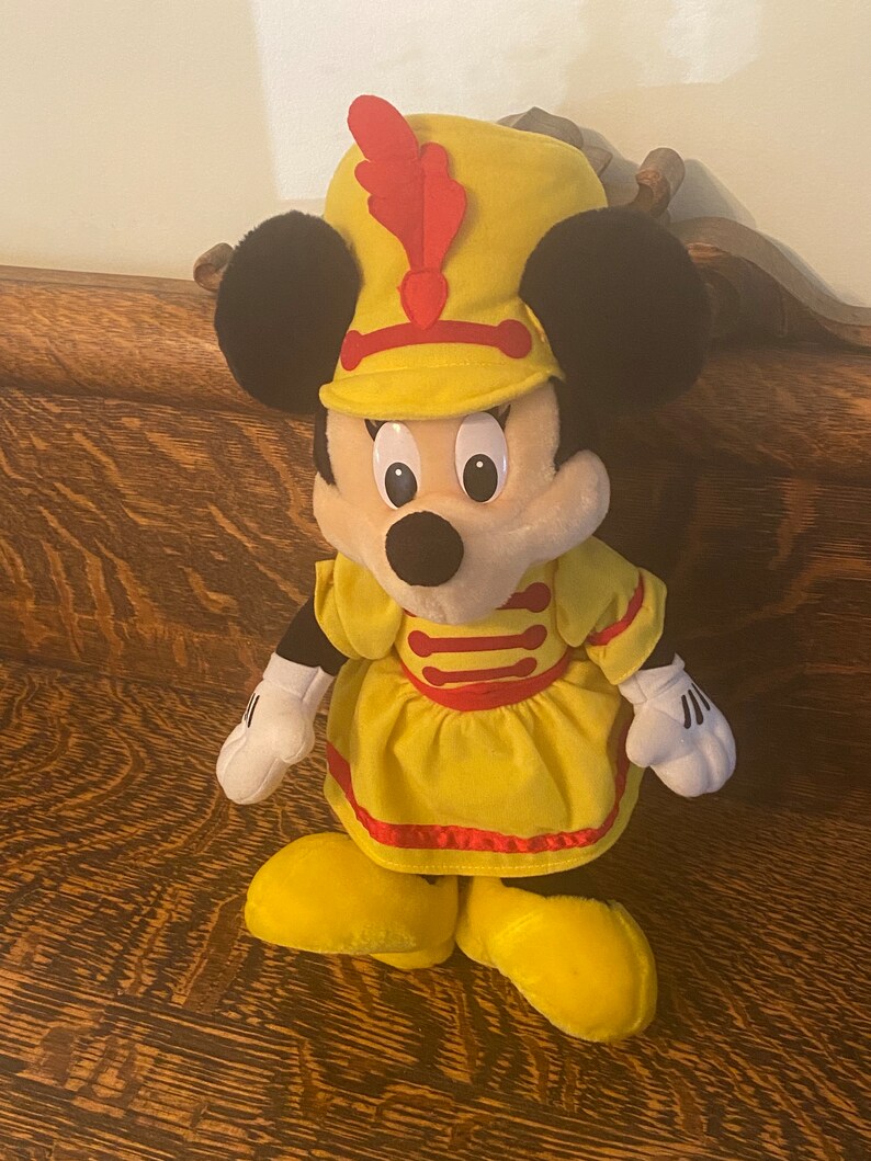 May include: A yellow plush toy of Minnie Mouse dressed as a drum major. She is wearing a yellow dress with red trim and a yellow hat with a red feather.