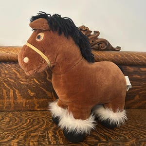 May include: A brown plush horse toy with a black mane and tail, and white fluffy fur around the hooves. The horse has a yellow bridle and large, round eyes. The toy is standing on a wooden surface.