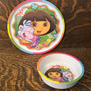 May include: A Dora the Explorer themed plate and bowl set. The plate and bowl have a white background with a colorful border. The plate features Dora and a monkey hugging. The bowl has Dora and the monkey with the word "Dora".