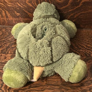May include: A green, turtle-shaped soft toy with a fluffy texture. It features a light tan beak and green flippers. The toy is positioned on a wooden surface.