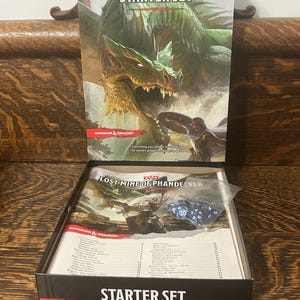 May include: A Dungeons & Dragons Starter Set box featuring a green dragon illustration on the lid. The open box contains the Lost Mine of Phandelver rulebook and a small clear plastic bag filled with blue polyhedral gaming dice for roleplaying.