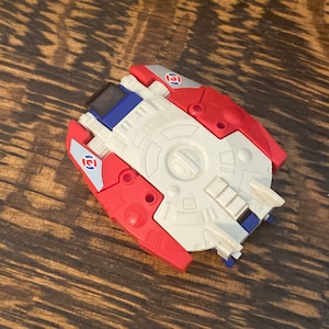 May include: A white, red, and blue plastic toy with a circular design and a small, rectangular window. The toy has a red and white design on the side and a blue and white design on the top.