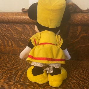 May include: A yellow plush toy dressed as a conductor with a black hat, red sash, and white lace trim. The toy is standing on a wooden surface.