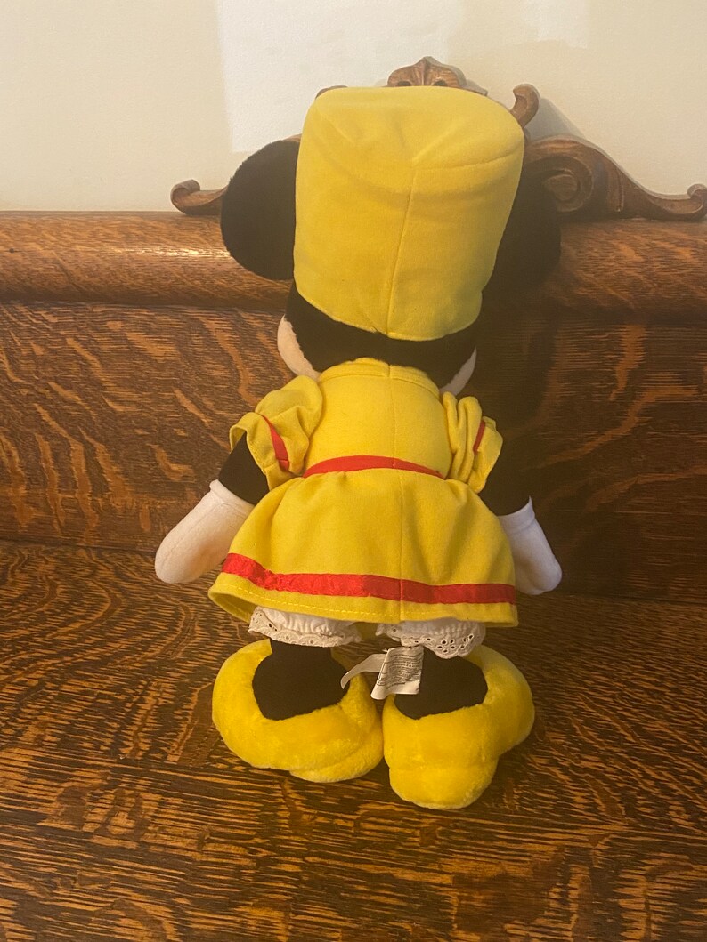 May include: A yellow plush toy dressed as a conductor with a black hat, red sash, and white lace trim. The toy is standing on a wooden surface.