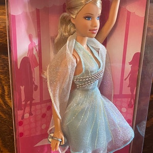 May include: A Barbie doll wearing a light blue dress with a silver sequined bodice and a sheer white shawl. The doll is holding a silver clutch purse. The doll is in a pink and white box with the words "Barbie Fashion" printed on the bottom.