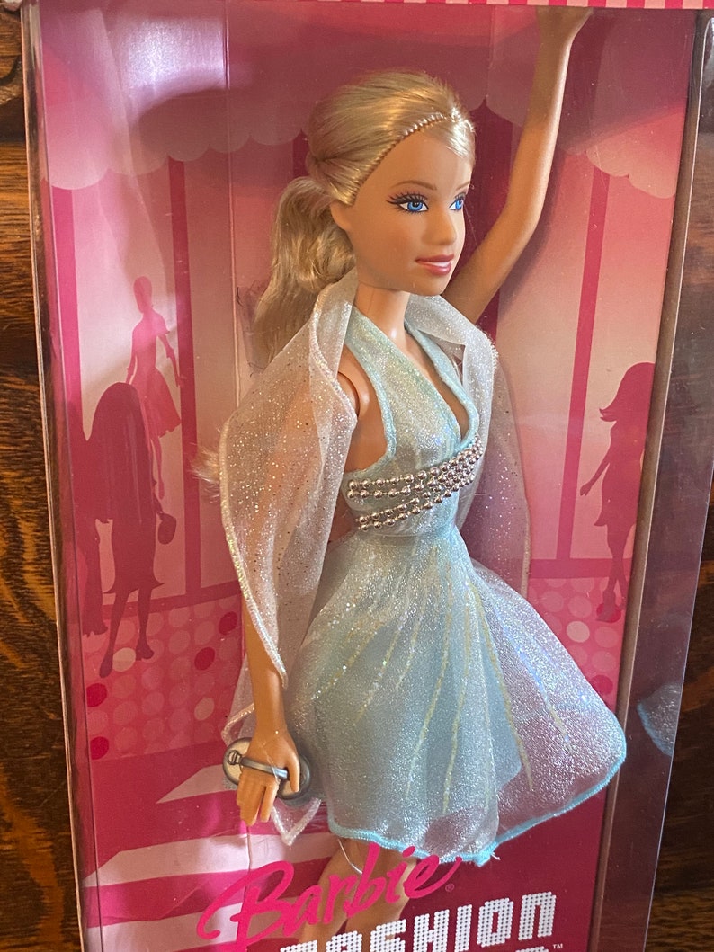 May include: A Barbie doll wearing a light blue dress with a silver sequined bodice and a sheer white shawl. The doll is holding a silver clutch purse. The doll is in a pink and white box with the words "Barbie Fashion" printed on the bottom.