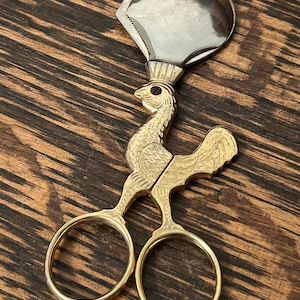 May include: A decorative scissor-style nutcracker with a rooster design. The top portion is silver-colored, while the body and handles are gold-toned. The scissor handles have circular finger holes. The nutcracker is set against a dark wood grain background.