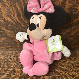 Op de afbeelding: Een Minnie Mouse knuffel met een zachte roze jurk en een bijpassende roze strik met witte stippen. Het personage heeft zwarte oren, een beige gezicht en witte handschoenen. Een groen label aan de zijkant toont het Disney Baby logo en tekst.