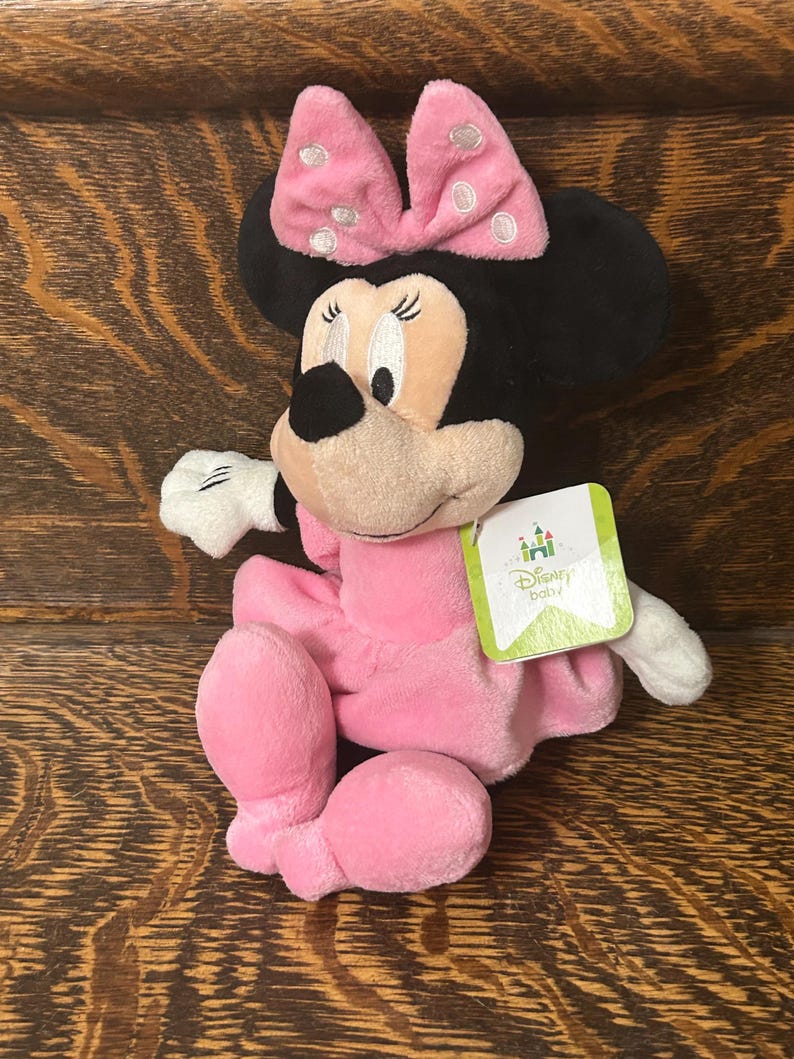 Op de afbeelding: Een Minnie Mouse knuffel met een zachte roze jurk en een bijpassende roze strik met witte stippen. Het personage heeft zwarte oren, een beige gezicht en witte handschoenen. Een groen label aan de zijkant toont het Disney Baby logo en tekst.