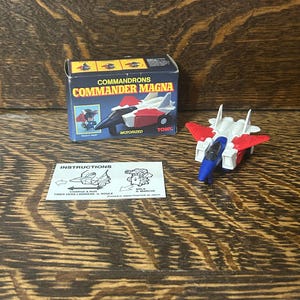 May include: A Tomy Commandrons Commander Magna motorized toy jet in white, red, and blue. The set includes a cardboard box with product imagery and text, plus a white instruction sheet with black line drawings showing how to operate the toy.