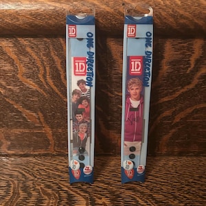 May include: Two One Direction merchandise packages. Each package is a vertical rectangle with blue top and bottom sections. The packages feature images of the band members and the text "ONE DIRECTION" and "I ❤️ 1D".
