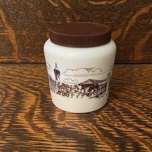 Op de afbeelding: Een witte keramische pot met een bruin deksel en een bruin-witte illustratie van een stadsscène. De illustratie bevat een kerk, een brug en mensen die lopen.