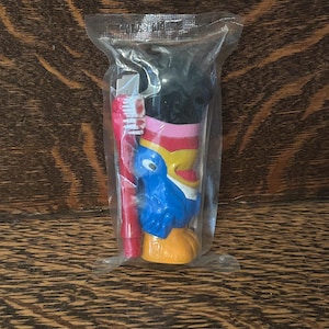 Puede incluir: Un paquete de plástico sellado que contiene un cepillo de dientes rojo y un soporte para cepillo de dientes temático. El soporte presenta un diseño de pájaro azul con detalles en amarillo, rosa y negro. El cepillo de dientes tiene cerdas blancas.