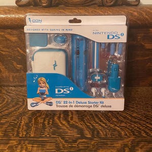 May include: A DSi 22-in-1 Deluxe Starter Kit in a clear plastic package. The kit includes a white and blue carrying case, a car charger, earbuds, and other accessories. The packaging features the Nintendo DS logo and the text "DESIGNED WITH GAMING IN MIND."