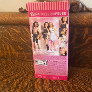 May include: A pink and white box with the words "Barbie Fashion Fever" on the front. The box features images of three Barbie dolls wearing different outfits. The text on the back of the box reads "Barbie - Doll/Poup&eacute;e/Puppe/Poppen/Mu&ntilde;eca/Bambola/Laleczka/Boneca/ドール - Stylish + Tons of Style! + School + Clothes + Shoes! + Get ready to rock!"