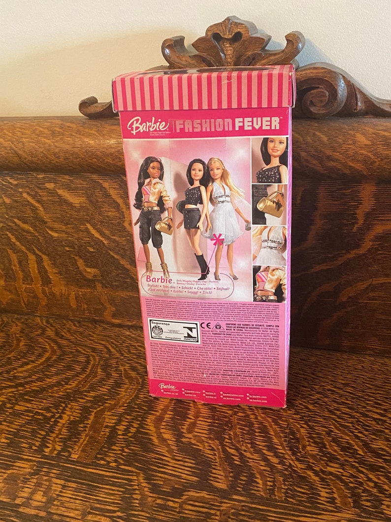 May include: A pink and white box with the words "Barbie Fashion Fever" on the front. The box features images of three Barbie dolls wearing different outfits. The text on the back of the box reads "Barbie - Doll/Poup&eacute;e/Puppe/Poppen/Mu&ntilde;eca/Bambola/Laleczka/Boneca/ドール - Stylish + Tons of Style! + School + Clothes + Shoes! + Get ready to rock!"
