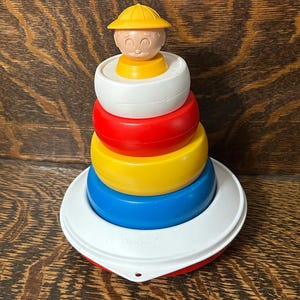 May include: A vintage stacking toy featuring a yellow-hatted figure atop a series of colorful rings. The rings are white, red, yellow, and blue, and sit on a white and red base. The toy is made of plastic.