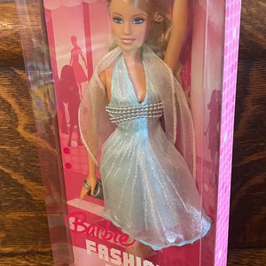 May include: A Barbie doll wearing a light blue sparkly dress with a silver beaded bodice. The doll is packaged in a pink box with the words "Barbie Fashion Fever" printed on it.