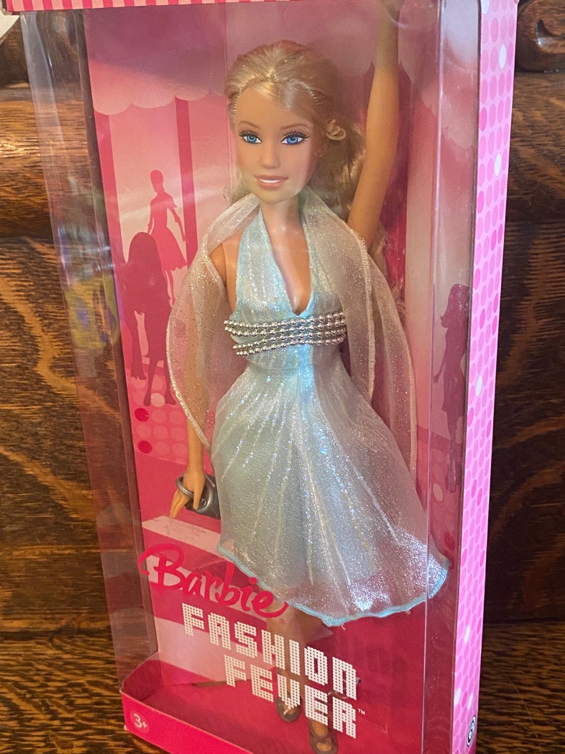 May include: A Barbie doll wearing a light blue sparkly dress with a silver beaded bodice. The doll is packaged in a pink box with the words "Barbie Fashion Fever" printed on it.