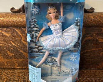 May include: A Barbie Snowflake doll in a collector's edition box. The doll is dressed in a white and light blue ballet costume with a tiara. The box features winter scenery and the text "Barbie Snowflake" and "The Nutcracker".