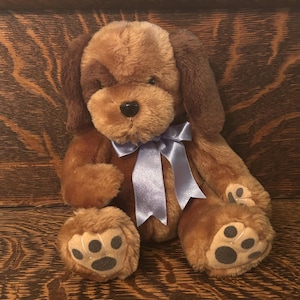 May include: A plush brown dog with floppy ears and a light blue ribbon around its neck. The stuffed animal has dark brown spots on its paws and a black nose. The dog is sitting on a wooden surface.