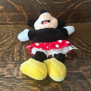 Pu&ograve; includere: Una bambola di peluche di Minnie Mouse, con un corpo nero, guanti bianchi e scarpe gialle. La bambola indossa una gonna rossa a pois bianchi e un bordo bianco a balze. Il viso della bambola &egrave; bianco con orecchie nere e bocca rosa.