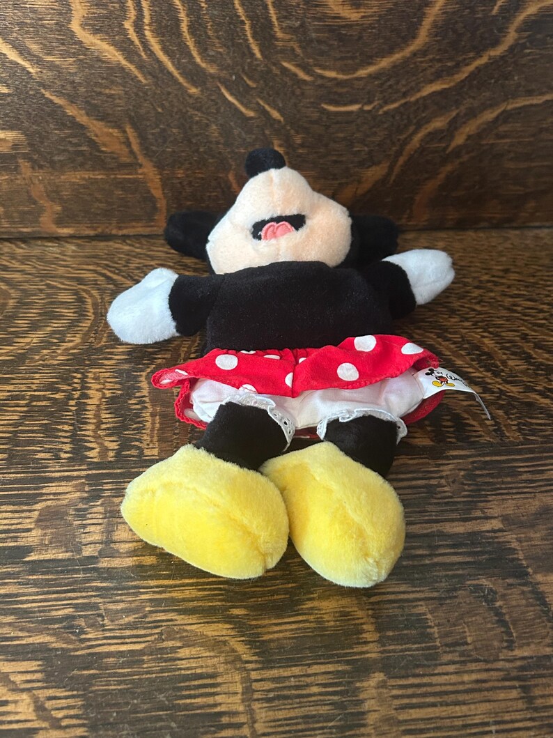 Pu&ograve; includere: Una bambola di peluche di Minnie Mouse, con un corpo nero, guanti bianchi e scarpe gialle. La bambola indossa una gonna rossa a pois bianchi e un bordo bianco a balze. Il viso della bambola &egrave; bianco con orecchie nere e bocca rosa.