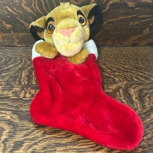 May include: A plush toy of a young lion character with tan fur, large brown eyes, and black-tipped ears, emerging from the top of a bright red Christmas stocking that features a soft white faux fur cuff along the upper edge of the opening.