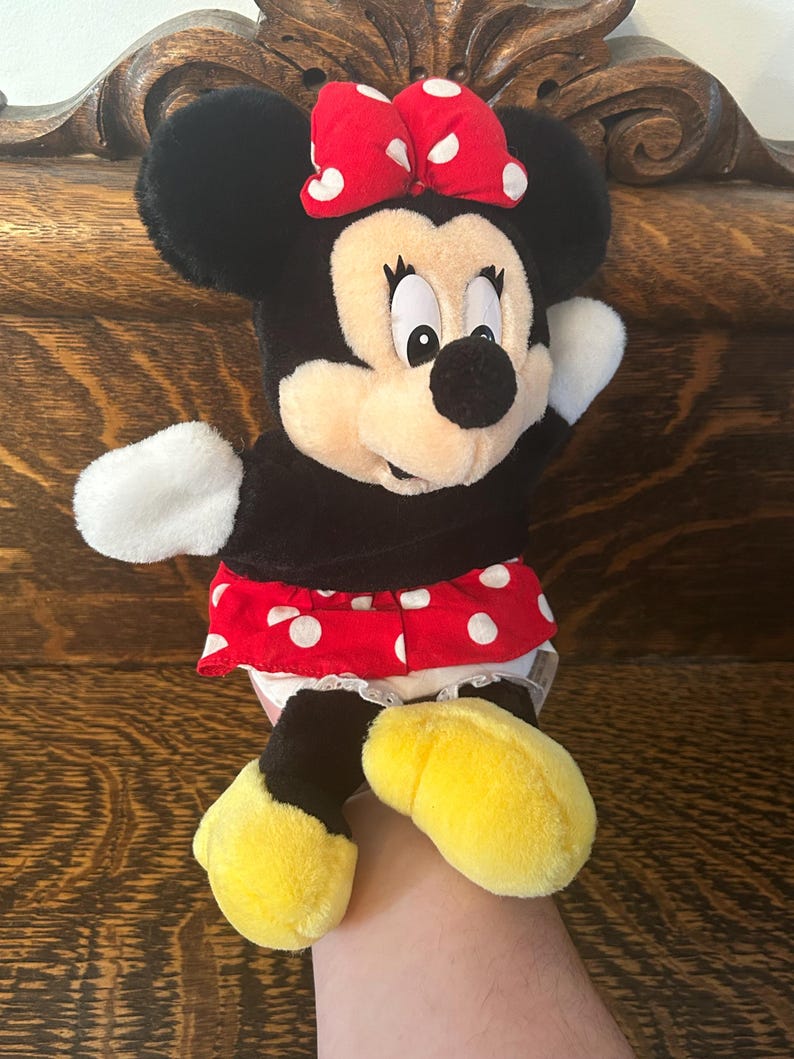 Pu&ograve; includere: Un peluche di Minnie Mouse. Il peluche ha un corpo nero, orecchie e un fiocco rosso a pois bianchi. Indossa una gonna rossa a pois bianchi e scarpe gialle. Il giocattolo &egrave; tenuto in mano da una persona.