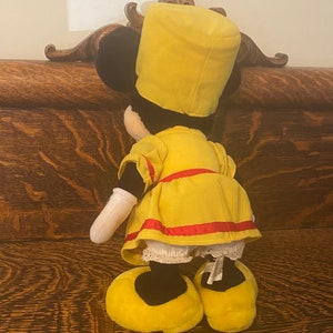 May include: A yellow plush toy with a black face and a yellow hat. The toy is wearing a yellow dress with a red sash and white lace trim. The toy is standing on a wooden surface.