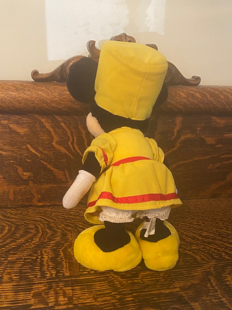 May include: A yellow plush toy with a black face and a yellow hat. The toy is wearing a yellow dress with a red sash and white lace trim. The toy is standing on a wooden surface.
