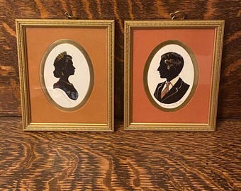 May include: Two framed silhouette portraits, one of a woman wearing a crown and the other of a man in a suit. The frames are gold with a brown mat and a white oval background.