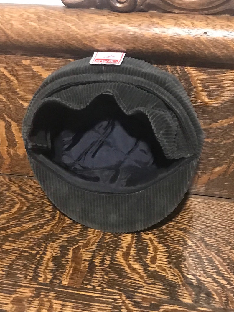 May include: A dark green corduroy newsboy cap with a red label that says "Kangol" on the front.