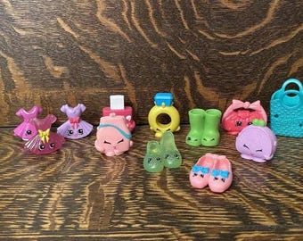 May include: A collection of ten colorful plastic toys featuring various characters and accessories. The toys are shaped like shoes, dresses, purses, and rings. The toys are all pink, green, blue, and yellow.