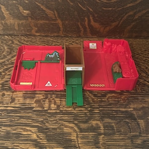 May include: A red Matchbox toy playset, open and unfolded, featuring a brown ramp, green and red components, and a small mountain scene. The playset includes a parking symbol and the Matchbox logo.