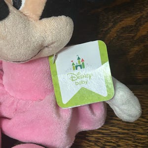 Op de afbeelding: Een Minnie Mouse knuffel met zwarte oren, een beige gezicht en een zachte roze jurk. Een wit label aan de zijkant toont een groen kasteelpictogram boven de tekst Disney baby, met een lichtgroene rand en subtiele cirkelvormige patronen.