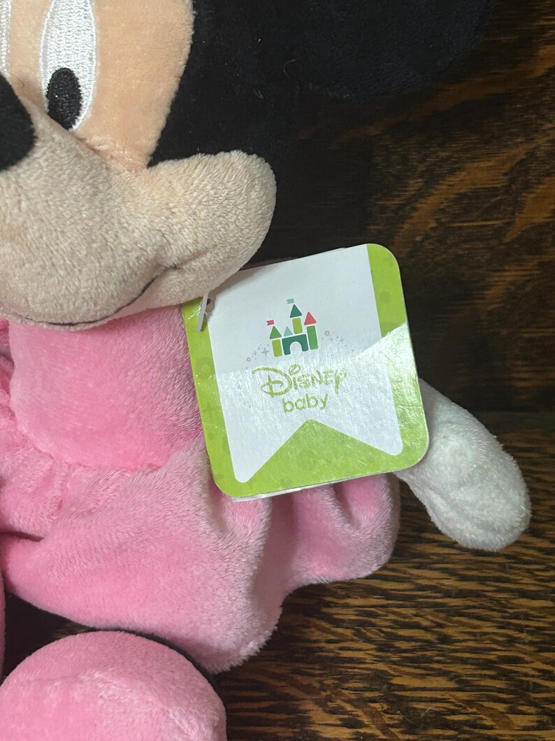 Op de afbeelding: Een Minnie Mouse knuffel met zwarte oren, een beige gezicht en een zachte roze jurk. Een wit label aan de zijkant toont een groen kasteelpictogram boven de tekst Disney baby, met een lichtgroene rand en subtiele cirkelvormige patronen.