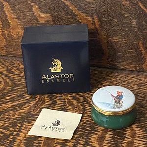 May include: A green and white enamel trinket box with a gold lid. The lid features a painting of a person fishing. The box is in its original packaging, which is a black box with a gold unicorn logo and the text "Alaster Enamels".