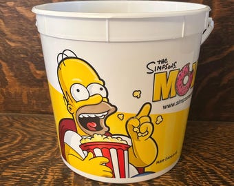 May include: White plastic popcorn bucket featuring Homer Simpson from The Simpsons cartoon. The bucket has a yellow and white design with Homer holding a popcorn container. The Simpsons logo and website are also visible.