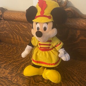 May include: A plush toy of Minnie Mouse dressed as a band member. She is wearing a yellow dress with red trim, a yellow hat with a red feather, and black shoes.