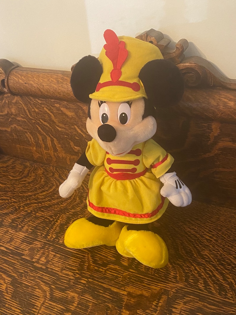 May include: A plush toy of Minnie Mouse dressed as a band member. She is wearing a yellow dress with red trim, a yellow hat with a red feather, and black shoes.