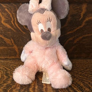 May include: A plush Minnie Mouse doll in pink, gray, and white. The doll has large gray ears, a pink bow, and a pink body. The doll is sitting on a wooden surface.