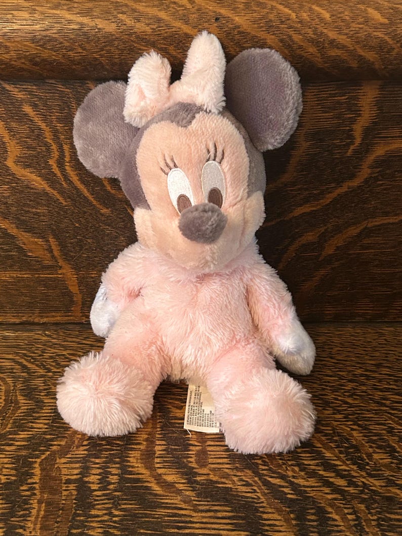 May include: A plush Minnie Mouse doll in pink, gray, and white. The doll has large gray ears, a pink bow, and a pink body. The doll is sitting on a wooden surface.